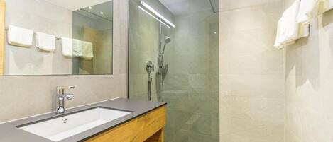 Un baño moderno con un espejo grande, un tocador de madera, ducha con mampara de vidrio y suelo de baldosas.