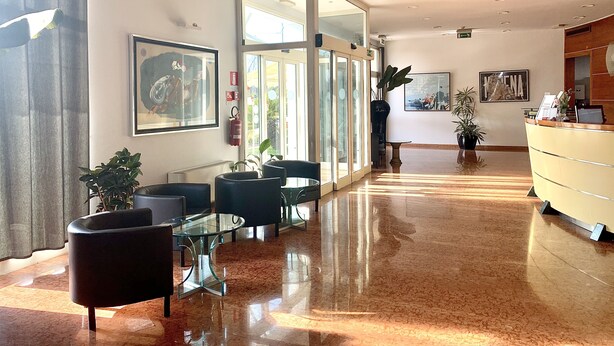 Eine Hotelhalle mit Rezeption, einer Sitzecke mit Stühlen und Tischen sowie einem großen Glasbalkon zur Außenwelt.