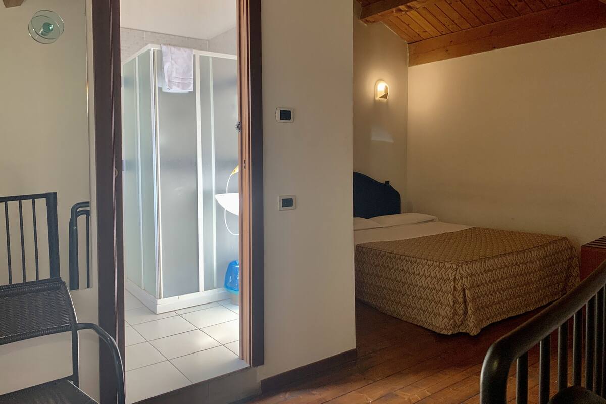 A bedroom with a bed, a chair, a wooden ceiling, and a bathroom visible through an open door.