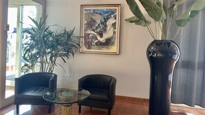 Eine Lobby mit Glastisch, zwei Ledersesseln, einer großen Topfpflanze und einem gerahmten Gemälde an der Wand.