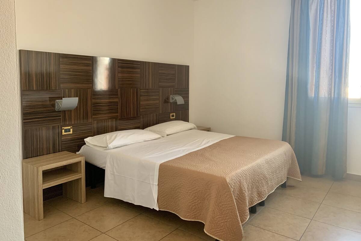 A bedroom with a wooden headboard, two bedside tables, a bed with white linens, a beige throw blanket, and a window with blue curtains.