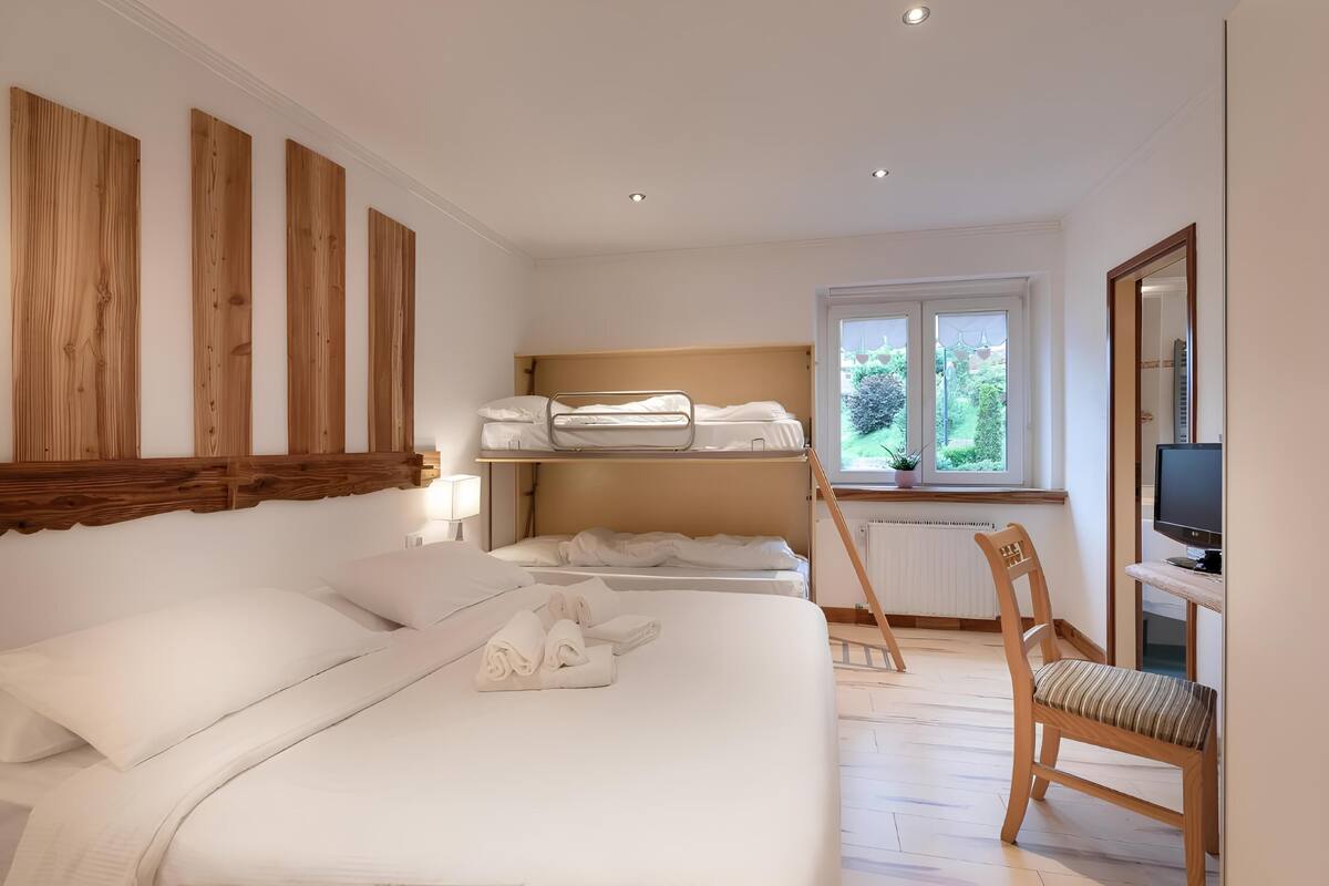 Hotelkamer met een groot bed, een kleiner bed op een verhoging, een stoel, een bureau met een computer en een raam met uitzicht op groen.