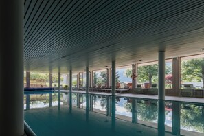 Une piscine intérieure avec des parois en verre transparent, des chaises longues et une vue sur des arbres et une clôture en bois.