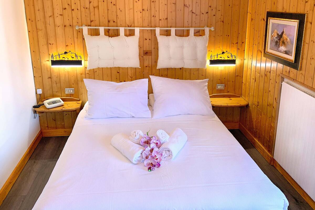 Een net opgemaakt bed met witte lakens en een handdoekboeket, houten wanden en bedlampjes.