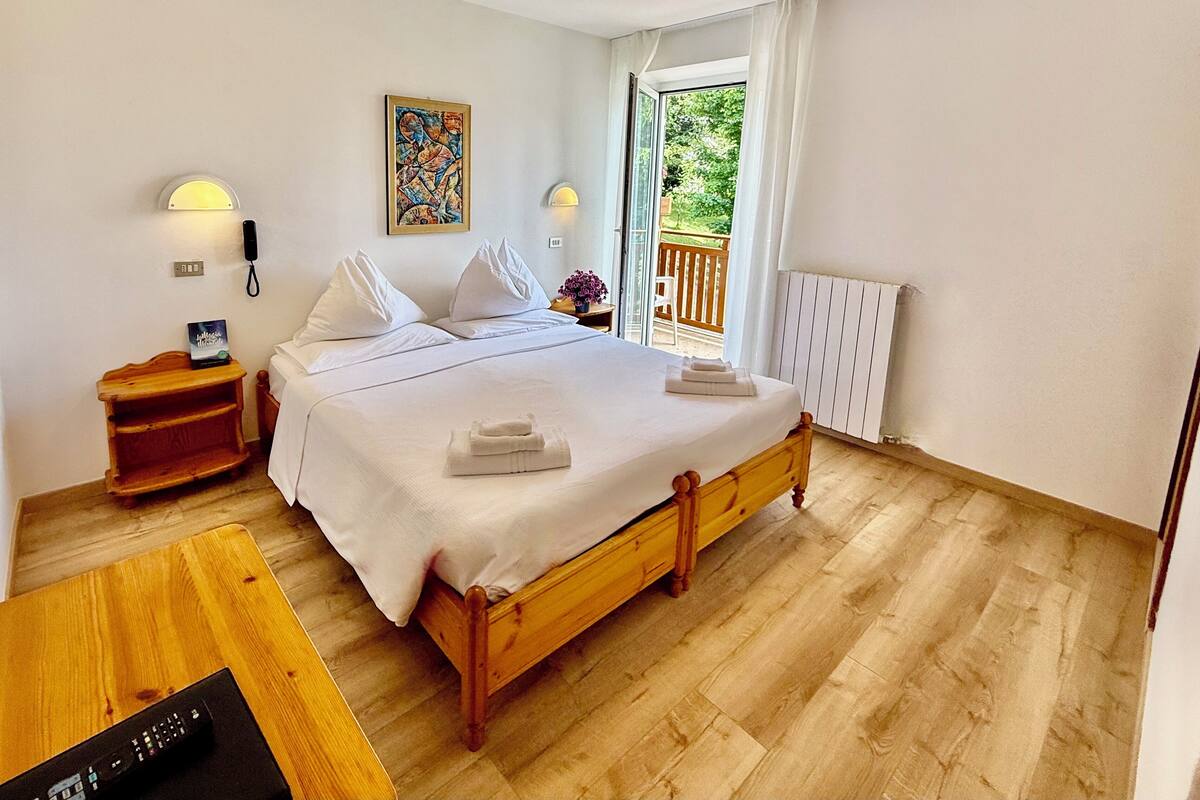 Een slaapkamer met een houten bed, nachtlampen, een nachtkastje, een houten tafel en uitzicht op de omgeving door een schuifdeur.