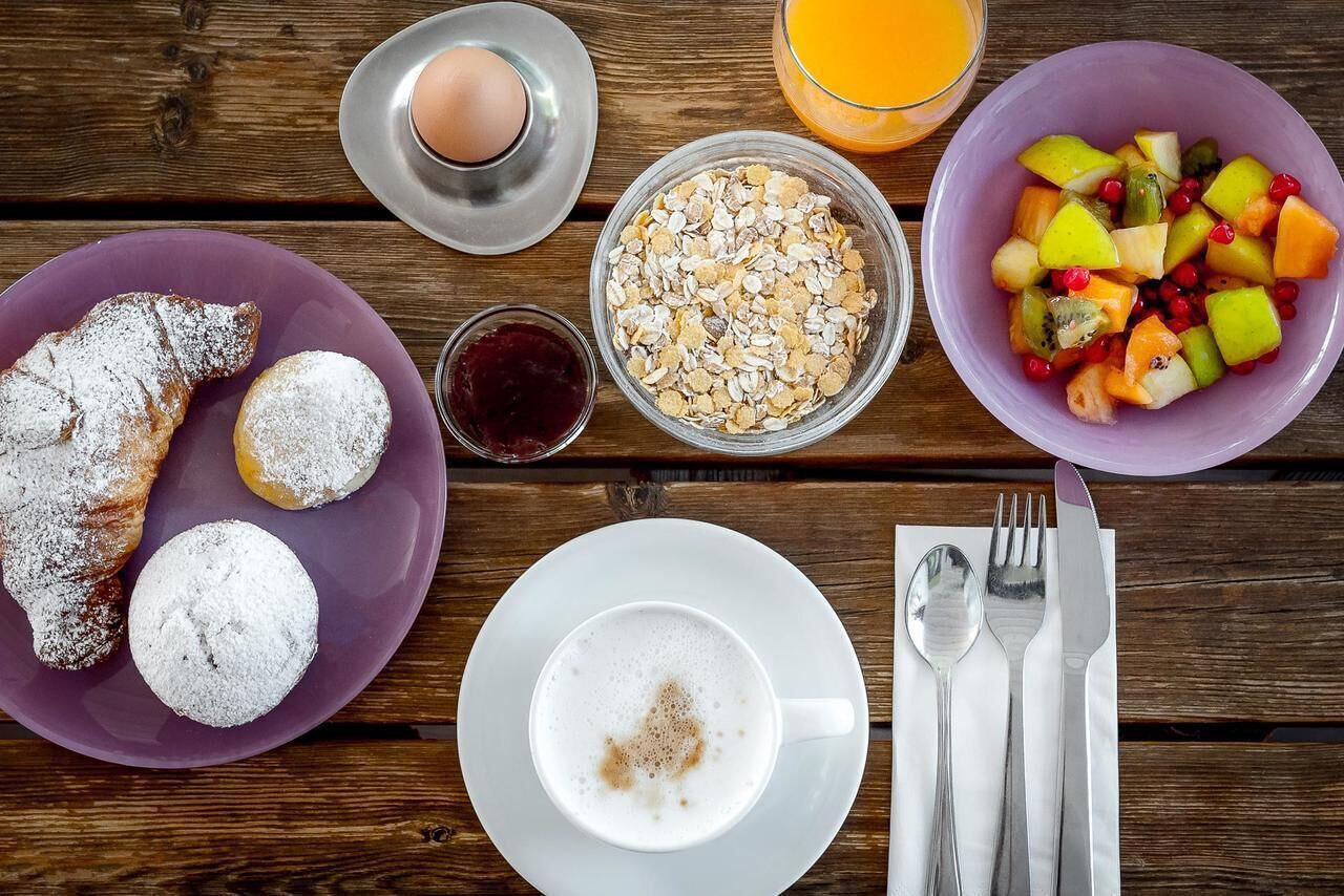 A breakfast setting with croissants, a bowl of cereal, a fruit salad, a glass of orange juice, a cup of coffee, and a plate with a boiled egg.