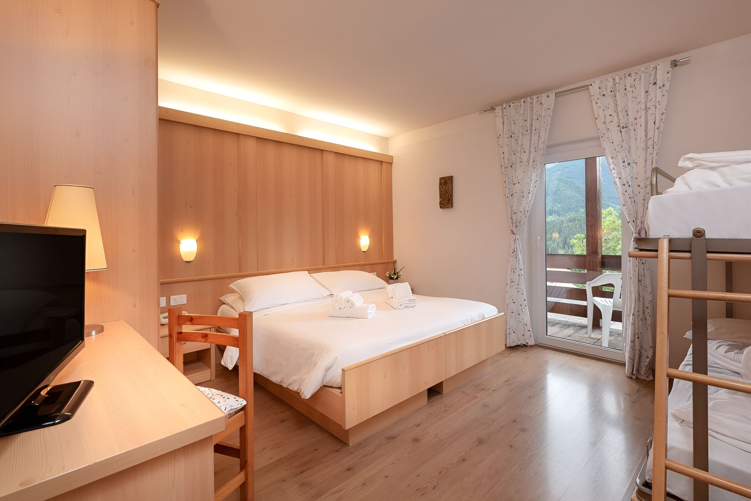A bedroom with a bed, a desk with a TV, a chair, and a balcony with a view.