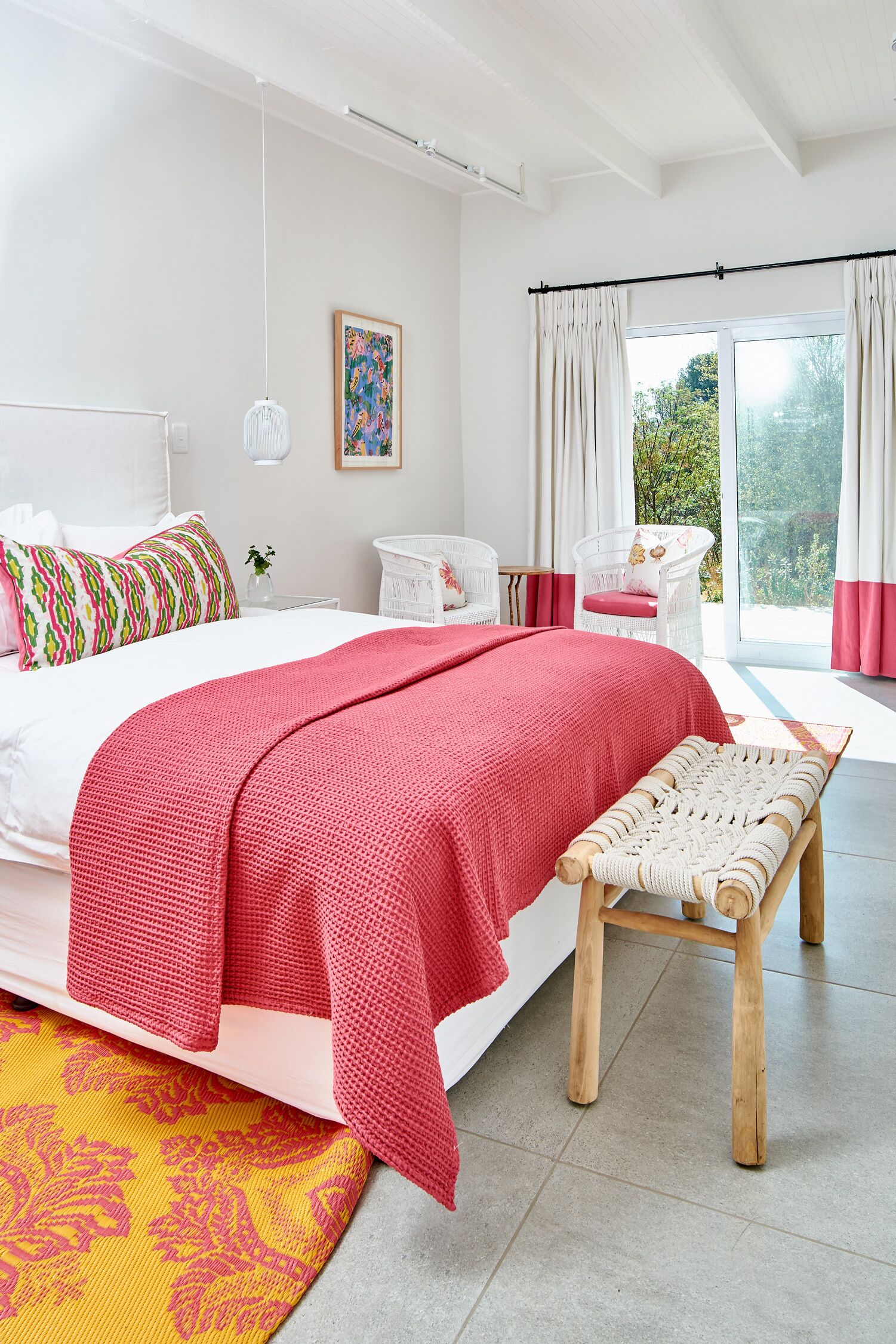 A modern bedroom with a large bed, a bench, a chair, and a view of greenery outside.