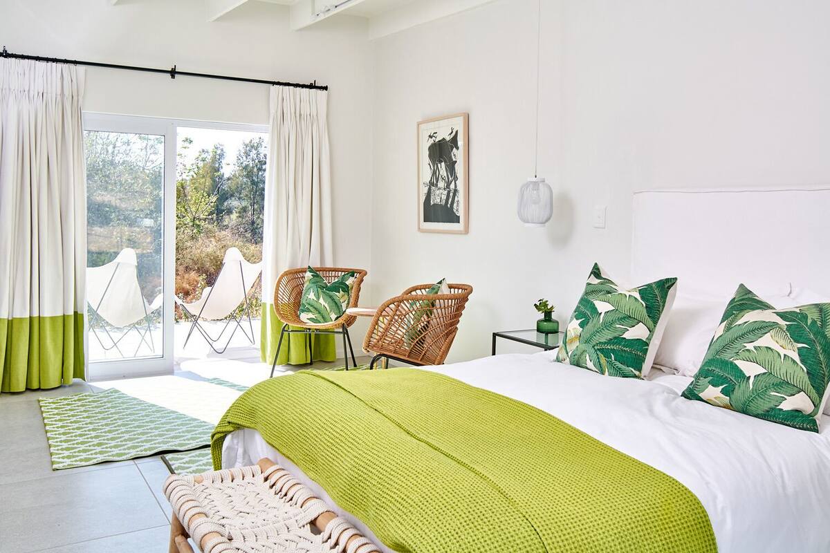 Un dormitorio con una cama, dos sillas de mimbre, una alfombra verde y vistas al exterior.