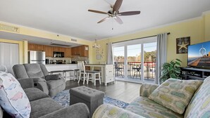 Ein Wohnzimmer mit grauen Sofas, einem Couchtisch und einem Fernseher. Im Hintergrund ist eine Küche mit Holzschränken und einem Deckenventilator zu sehen.