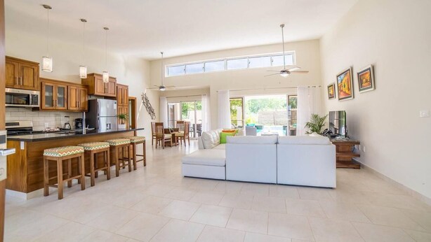 A spacious kitchen with wooden cabinets, a central island with bar stools, a dining area with a table and chairs, a large white sofa, and a ceiling fan. - Contemporary Design With Lots of big Windows (Sosúa)