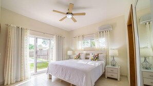 A bedroom with a large bed, a ceiling fan, a window with curtains, a bedside table with a lamp, and an air conditioning unit. - Contemporary Design With Lots of big Windows (Sosúa)