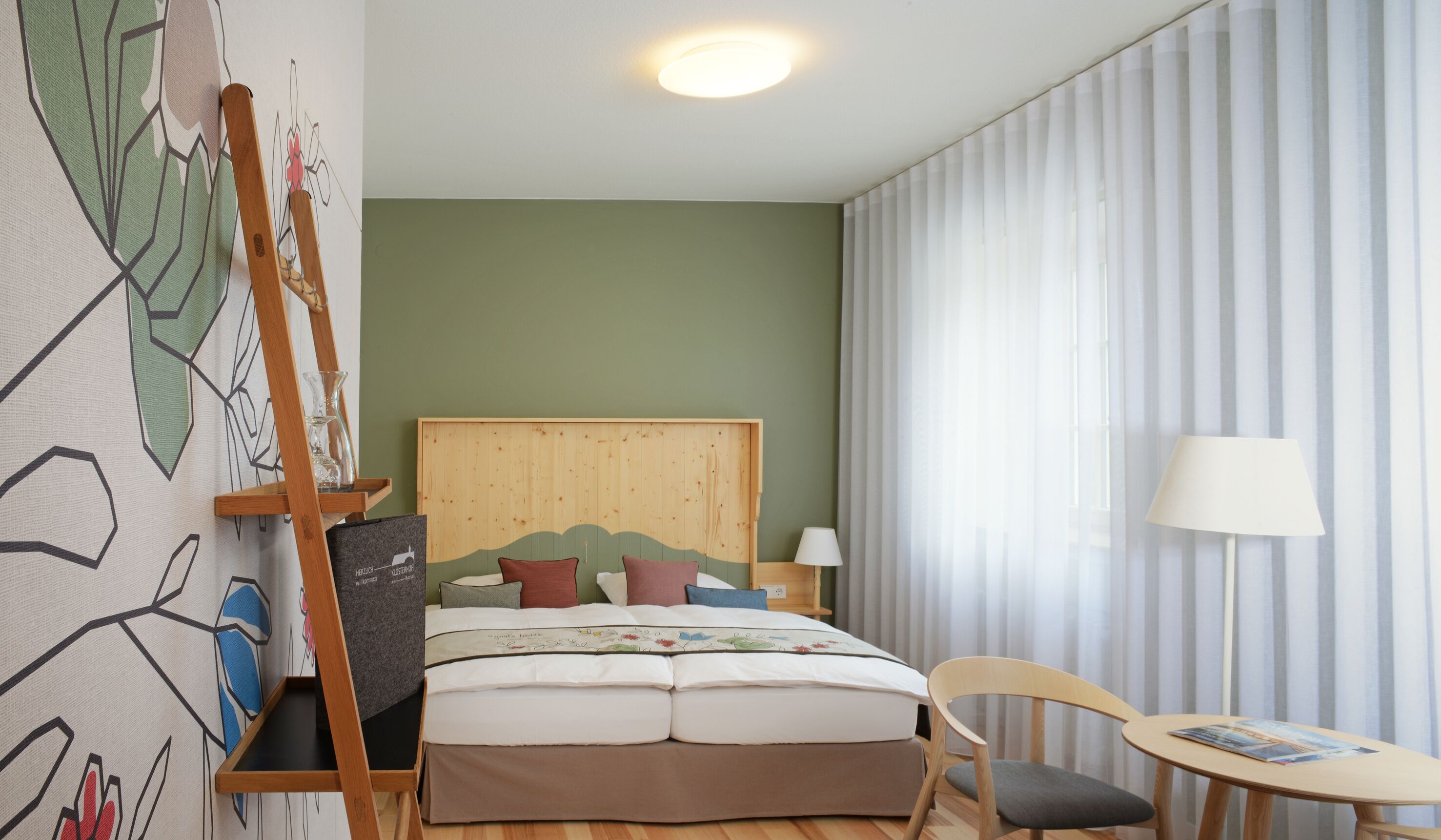 Una habitación de hotel con cabecera de madera, una cama con varias almohadas, una mesita de noche con lámpara, una silla y una pequeña mesa con una revista.