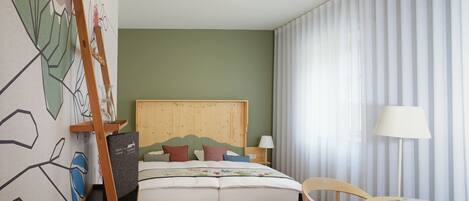 Ein Hotelzimmer mit Holz Kopfteil, einem Bett mit vielen Kissen, einem Nachttisch mit Lampe, einem Sessel und einem kleinen Tisch mit einer Zeitschrift.