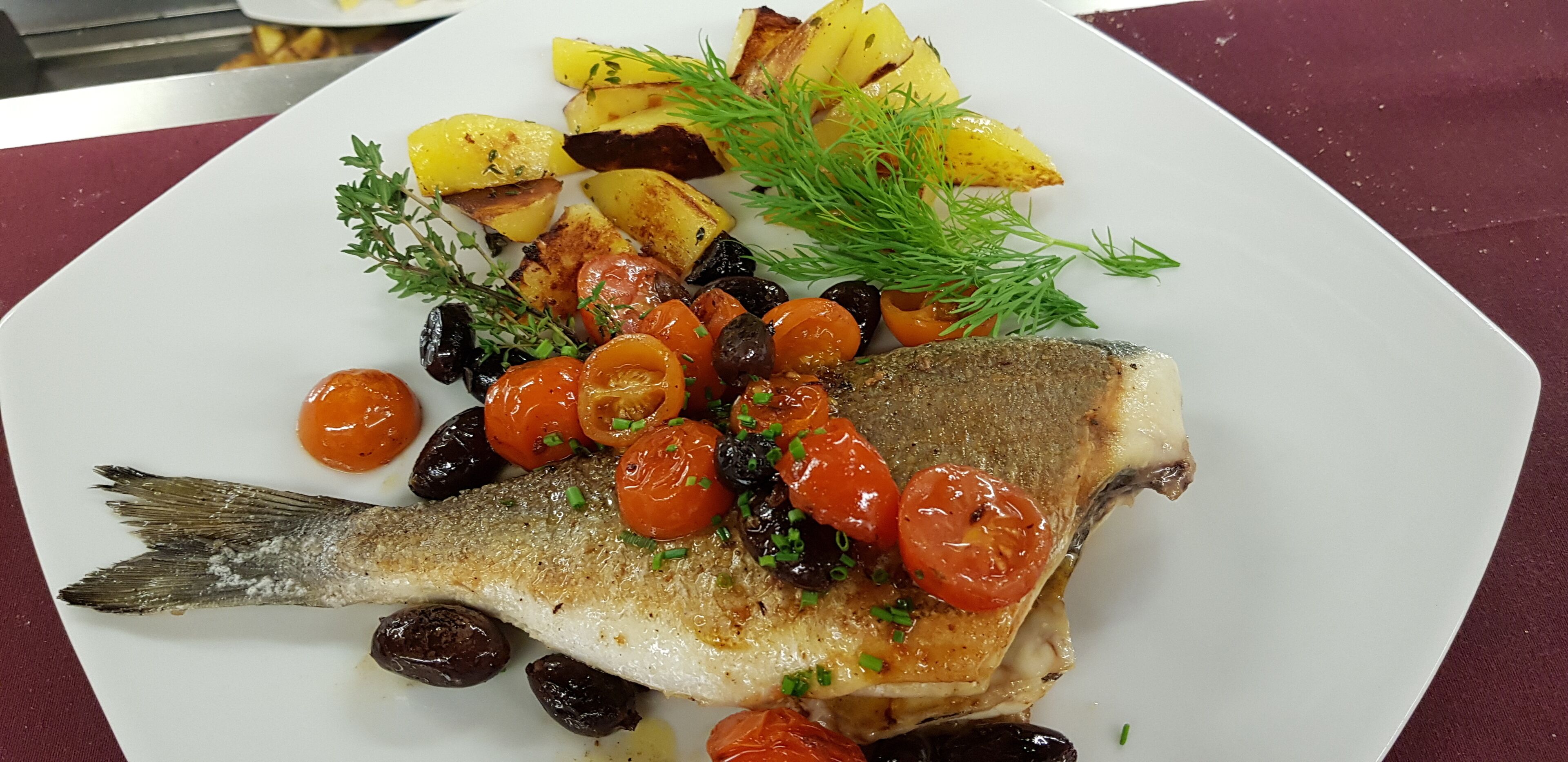 Une assiette blanche contenant un poisson grillé, des pommes de terre rôties et des tomates cerises, le tout garni d’herbes aromatiques.