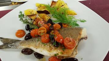 Une assiette blanche contenant un poisson grillé, des pommes de terre rôties et des tomates cerises, le tout garni d’herbes aromatiques.