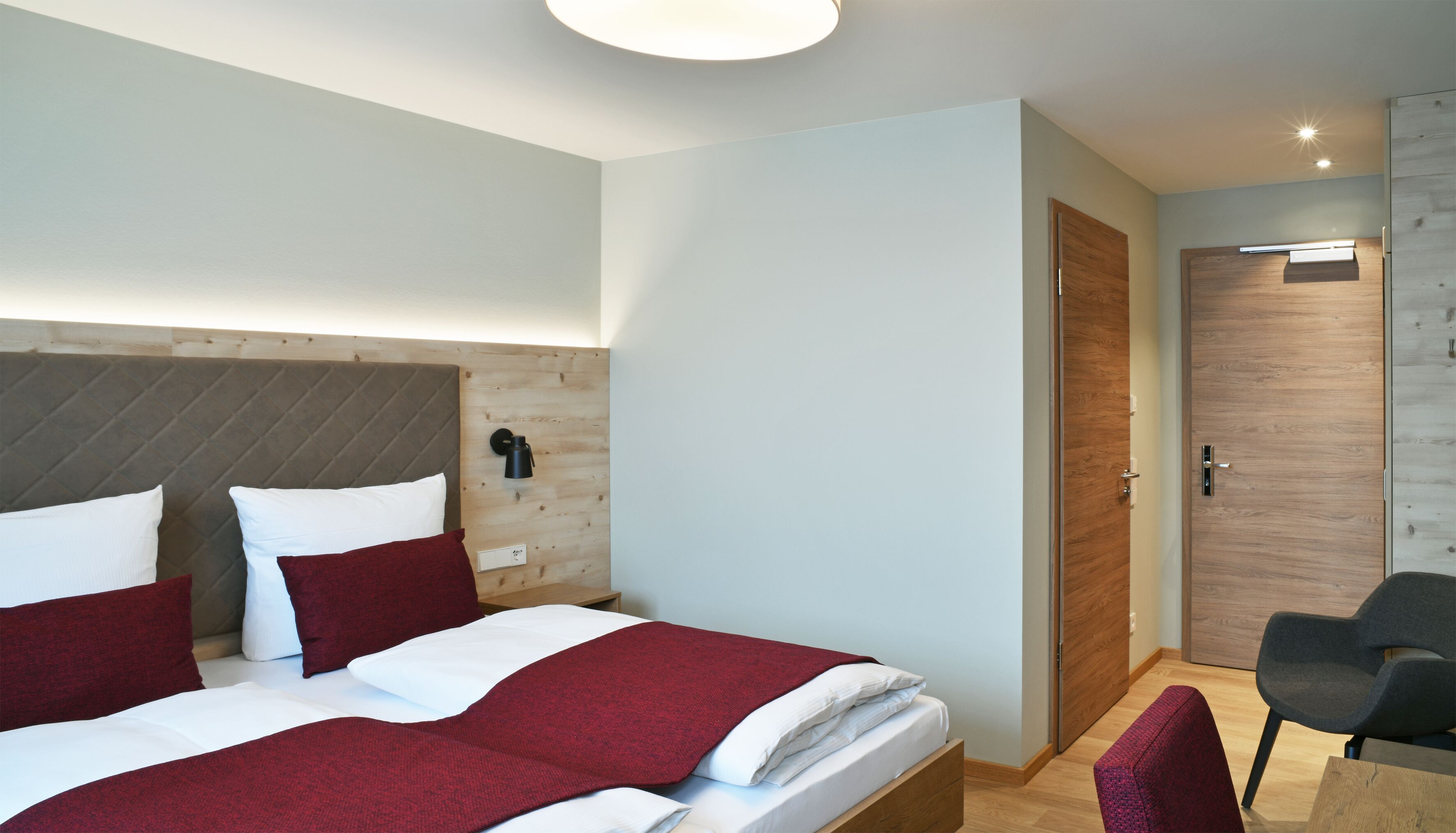 A modern hotel room with a large bed, a wooden headboard, a bedside table, a nightstand, a chair, and a wardrobe.