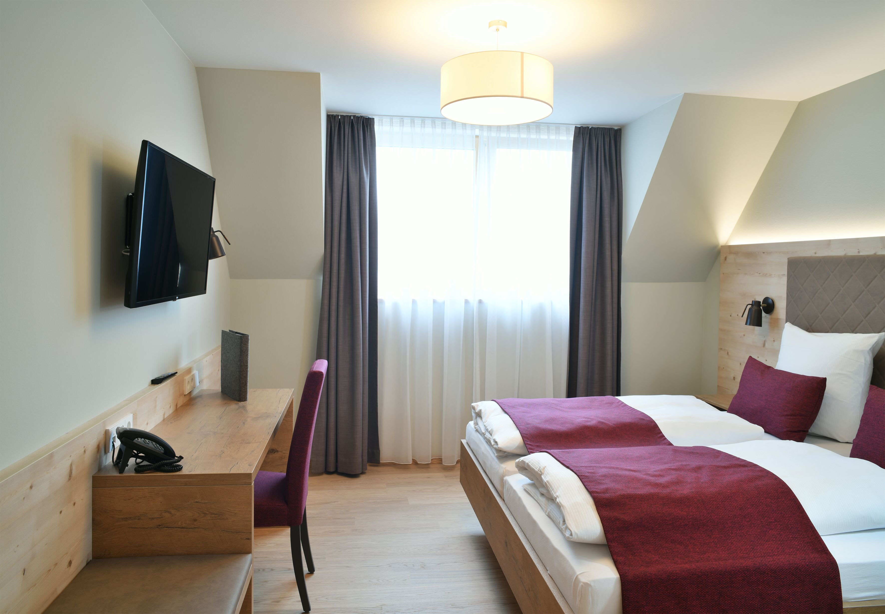 Una habitación de hotel con cama, escritorio con teléfono, una silla, televisor montado en la pared y una ventana con cortinas.