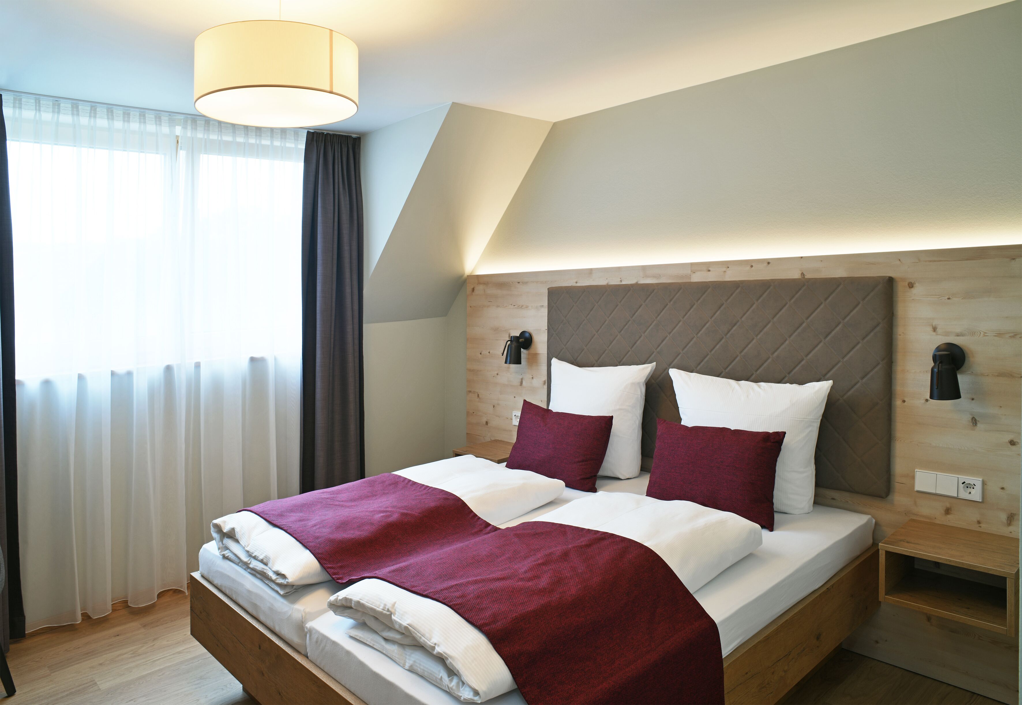 Een tweepersoonsbed met een houten hoofdbord, beddengoed in wit en rood, en een nachtkastje met een lamp.