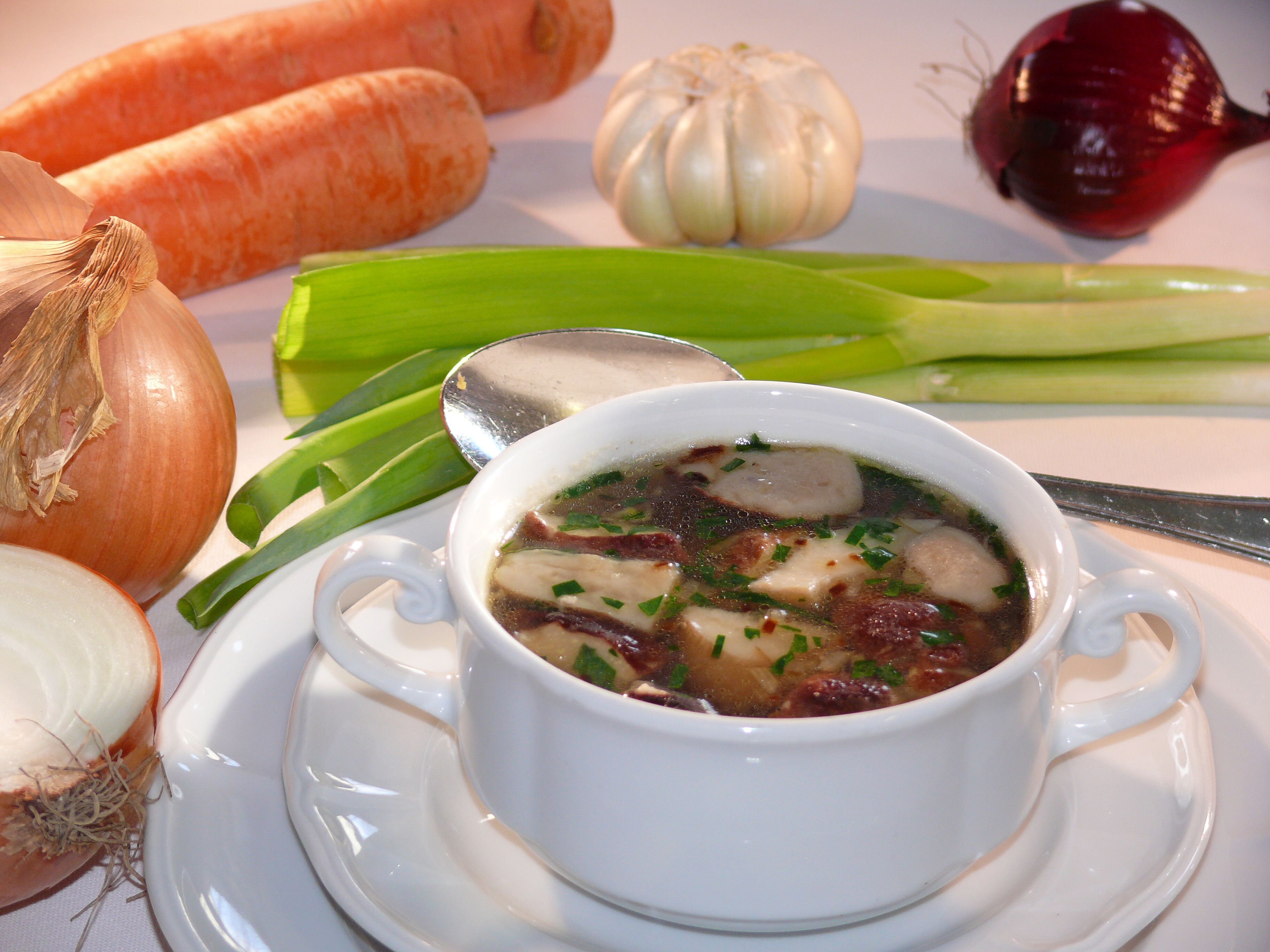 Un tazón de sopa con verduras y hierbas, rodeado de vegetales crudos como zanahorias, cebollas y ajo.