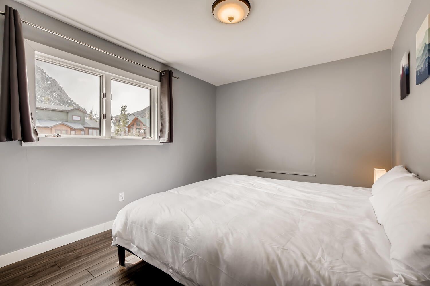 Ein Schlafzimmer mit einem großen Bett, einem Fenster mit Vorhängen, einem Nachttisch mit Lampe und Blick auf Häuser und Berge.