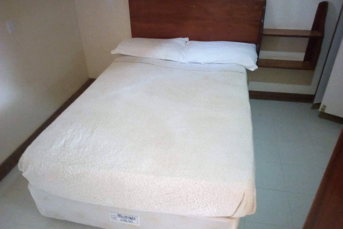 Ein kleines, ordentlich bezogenes Bett mit Holz Kopfteil und weißer Bettwäsche in einem schlichten Zimmer mit weißen Wänden und Fliesenboden.