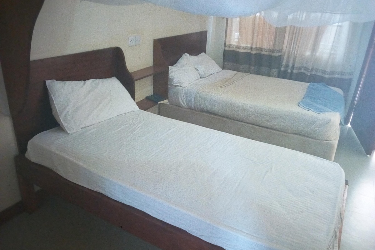 Ein Zimmer mit zwei Einzelbetten, Holz-Kopfteilen, weißer Bettwäsche, einem Nachttisch und einem Fenster mit durchsichtigen Vorhängen.