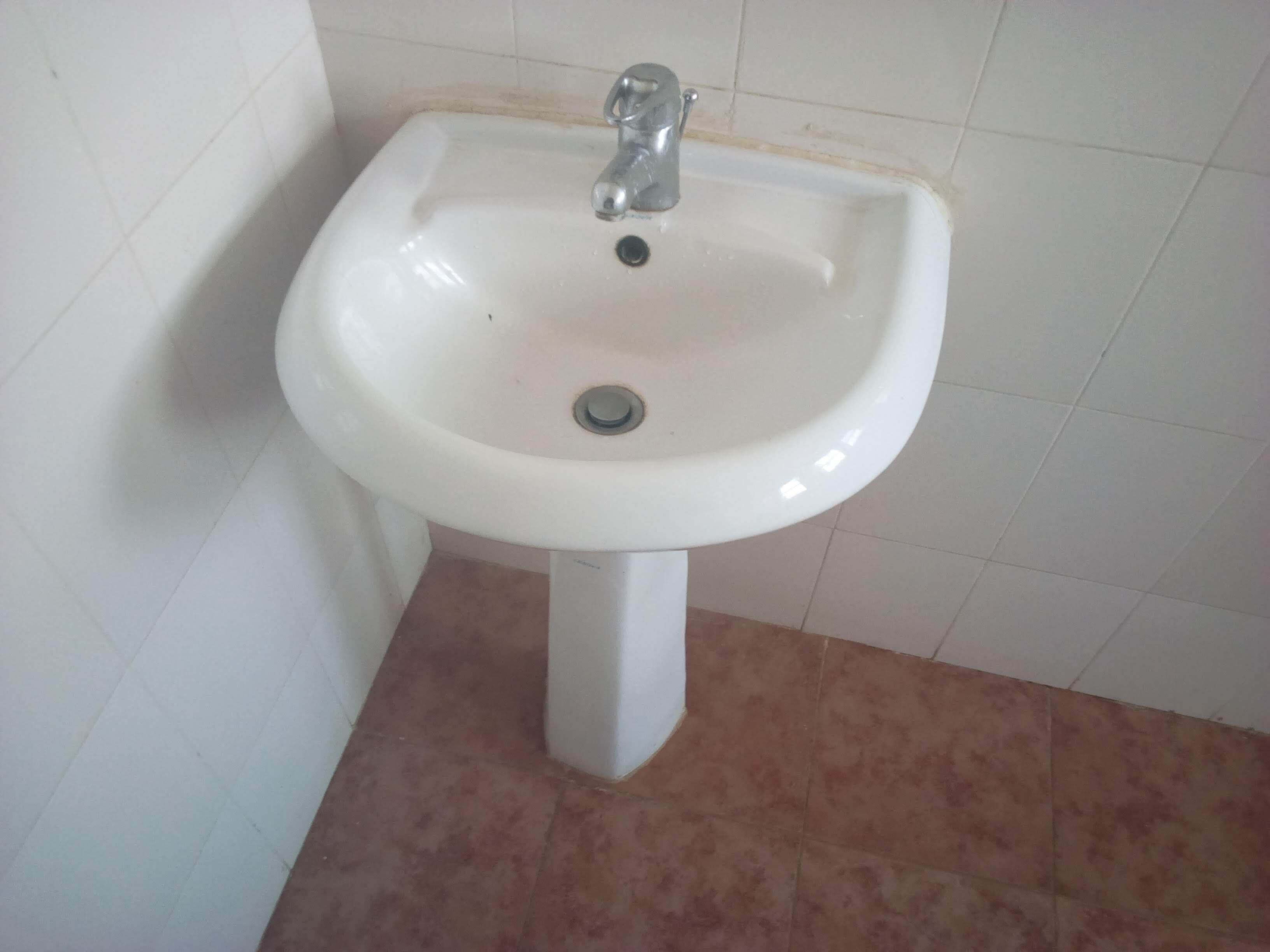 Un lavamanos blanco sobre pedestal con un solo grifo, frente a una pared y piso cubiertos de azulejos.
