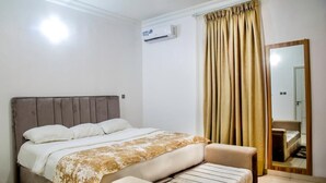 A bedroom with a bed, a bedside table, a chair, a large mirror, and a window with curtains. - Bella Residencia (Lekki)