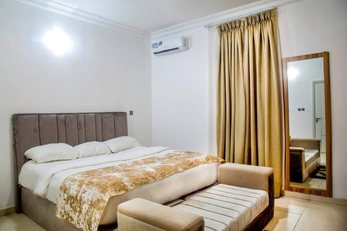 Quarto com cama, mesa de cabeceira, cadeira, espelho grande e janela com cortinas.
