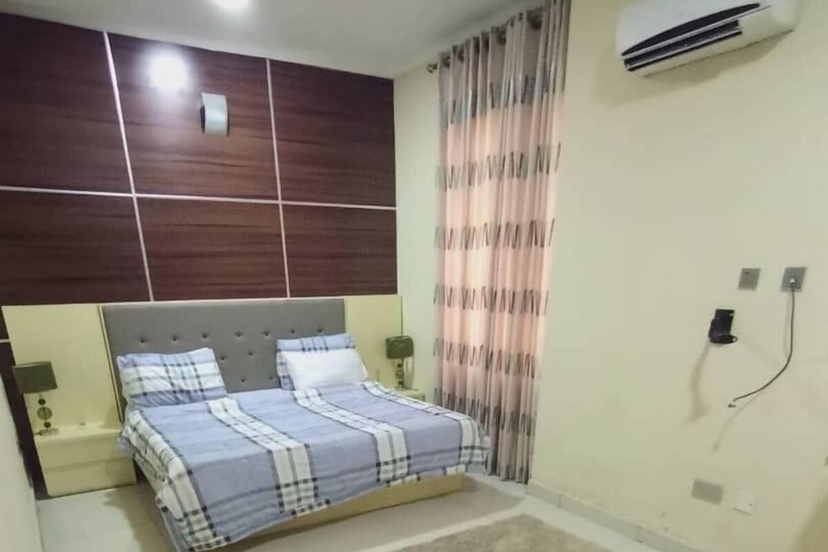 Quarto com cama, tapete, mesa de cabeceira, luminária, ar condicionado e cortina.