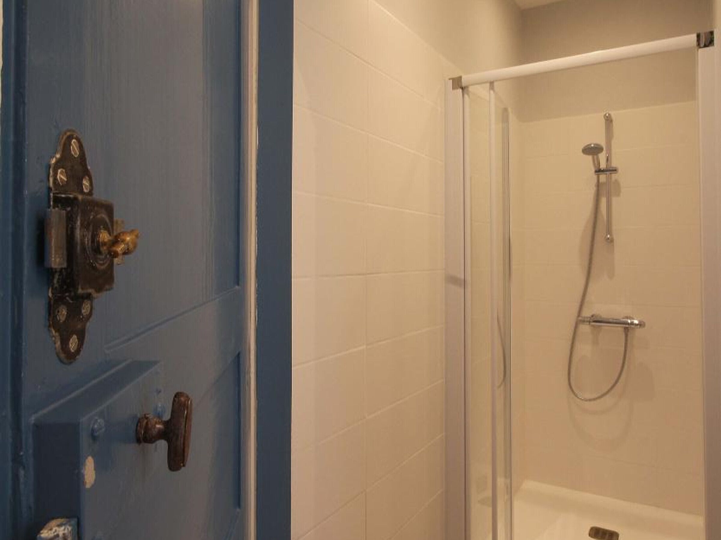 Un baño con ducha, una puerta azul con cerradura y una pared de azulejos blancos.