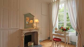 Une pièce avec une cheminée, un miroir, une table ornée d’un vase de fleurs et une fenêtre avec des rideaux.