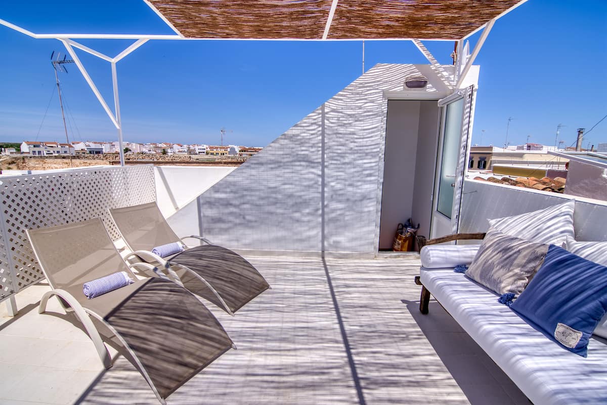 Una terraza exterior iluminada por el sol con sillones de descanso y un sofá, con vistas a una zona costera donde se divisan edificios a lo lejos.