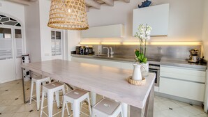 A modern kitchen with a central island, white bar stools, a hanging wicker light fixture, and a potted orchid. - Hotel Ca s'Arader - Adults Only (Ciutadella de Menorca)