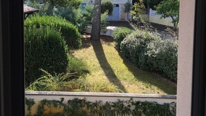 Blick auf einen Garten mit verschiedenen Bäumen und Sträuchern, ein Ziegeldach und ein Gebäude im Hintergrund.
