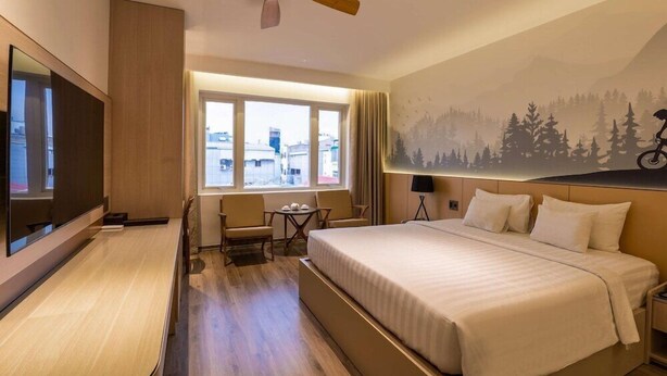 A hotel room with a bed, a desk, a chair, a TV, a ceiling fan, and a window with a view of buildings. - Triip Palago (Ho Chi Minh City)