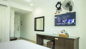A hotel room with a bed, a desk with a mirror, a wall-mounted TV displaying music, a clock, and a fan.