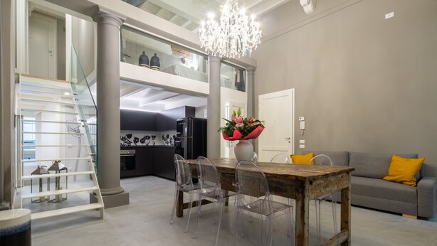 A spacious living area with a dining table, clear chairs, a chandelier, a grey sofa with a yellow pillow, and a staircase leading to an upper level. - Casa Velluti Deluxe by Mmega (Florence)