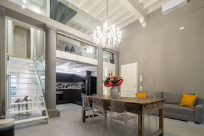 A spacious living area with a dining table, clear chairs, a chandelier, a grey sofa with a yellow pillow, and a staircase leading to an upper level.
