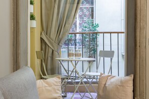 A balcony with a table and chairs, a view of the street, and a window with curtains.