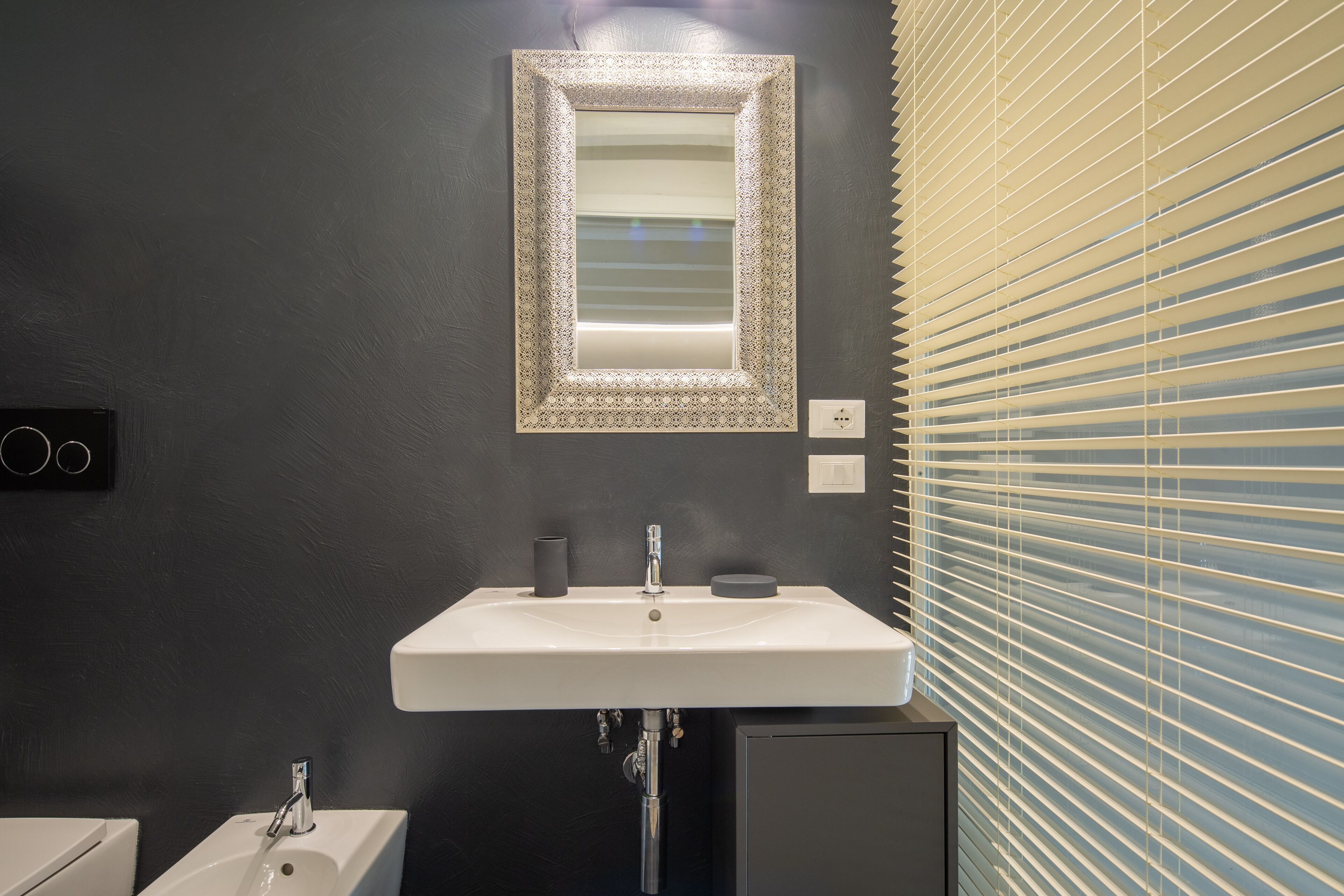 A bathroom with a white sink, a mirror with a decorative frame, and a window with blinds.