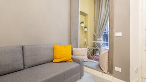 A grey sofa with a yellow pillow, a mirror reflecting a room with a chair and a table, and a door leading to another space. - Casa Velluti Deluxe by Mmega (Florence)