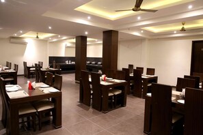 A restaurant with multiple dining tables and booths, ceiling fans, recessed lighting, and a kitchen area in the background. - The Raj Vilas (Agra)