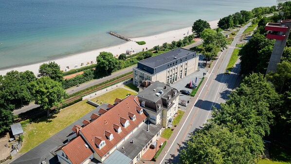 An aerial view of a coastal area with a beach, a modern building with a flat roof, and a road. - Hotel BeachSide (Eckernfoerde)