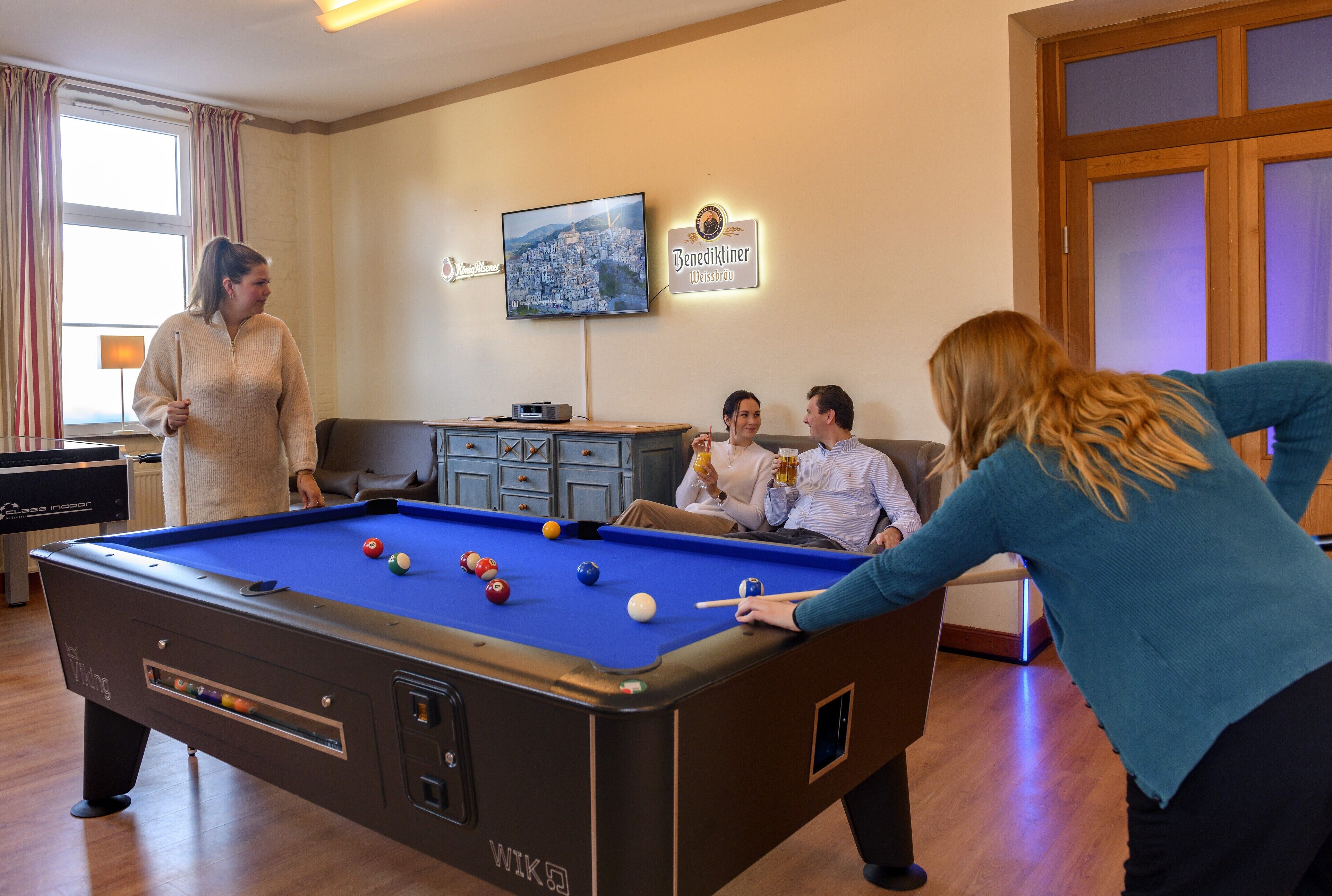 A pool table in a room with a cityscape TV, a sign reading "Benedikiner Wiesbräu," and a wooden cabinet.