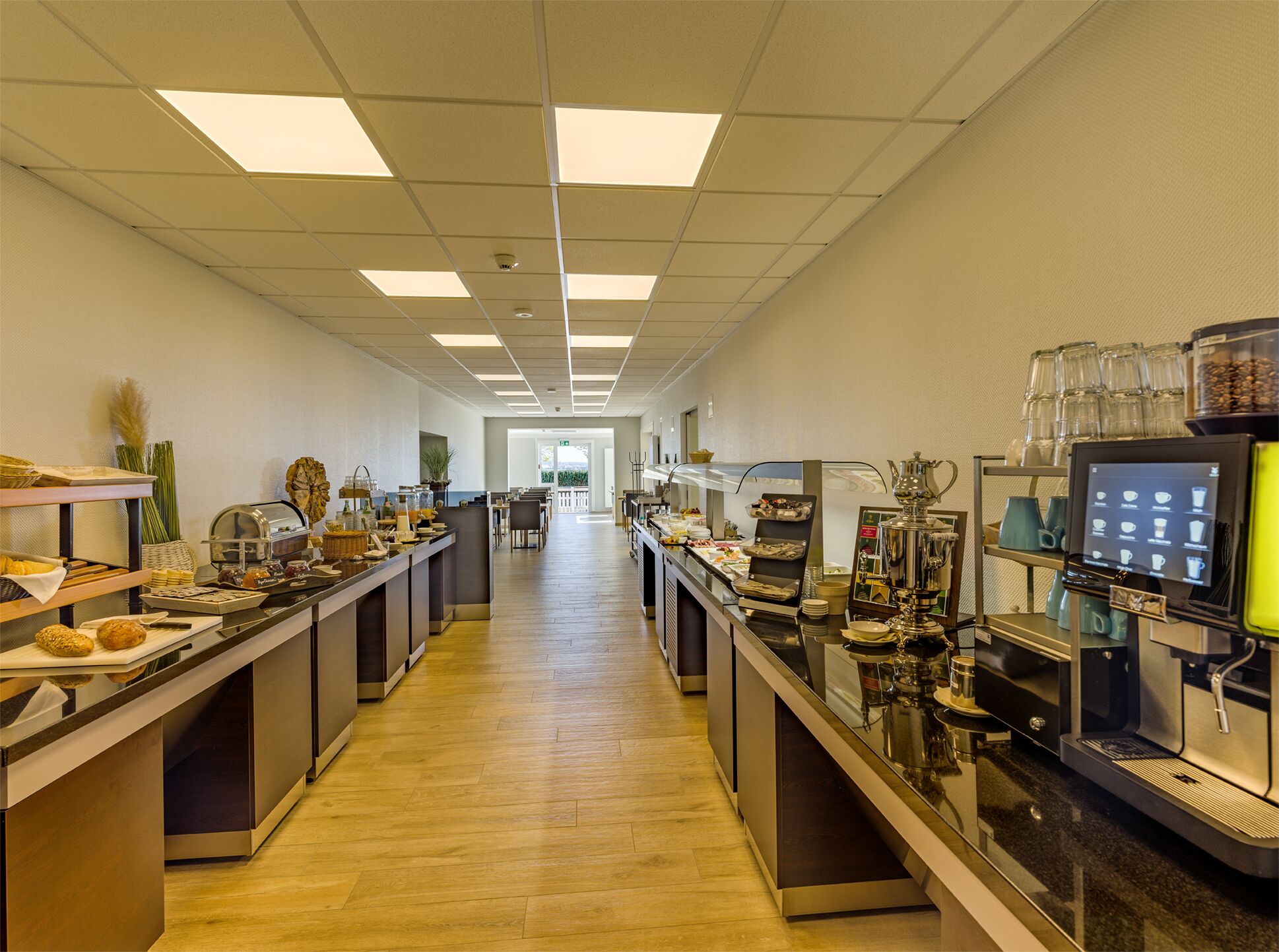 A long, well-lit kitchen area with multiple counters, coffee machines, and a display screen.