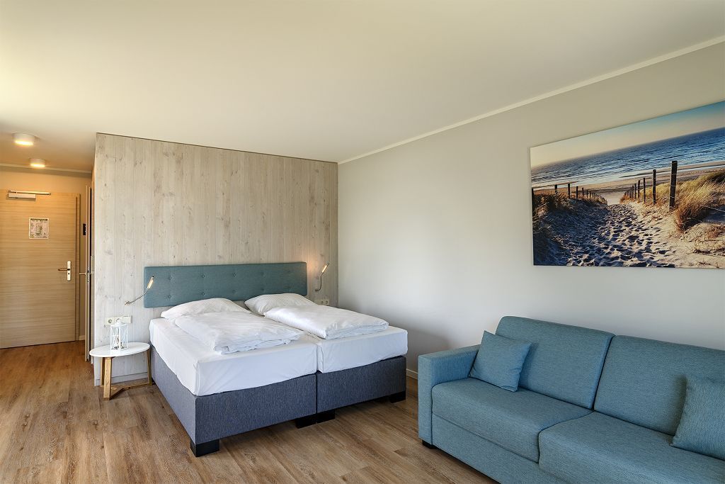 A bedroom with a bed, a blue sofa, a painting of a beach, and a wooden door.