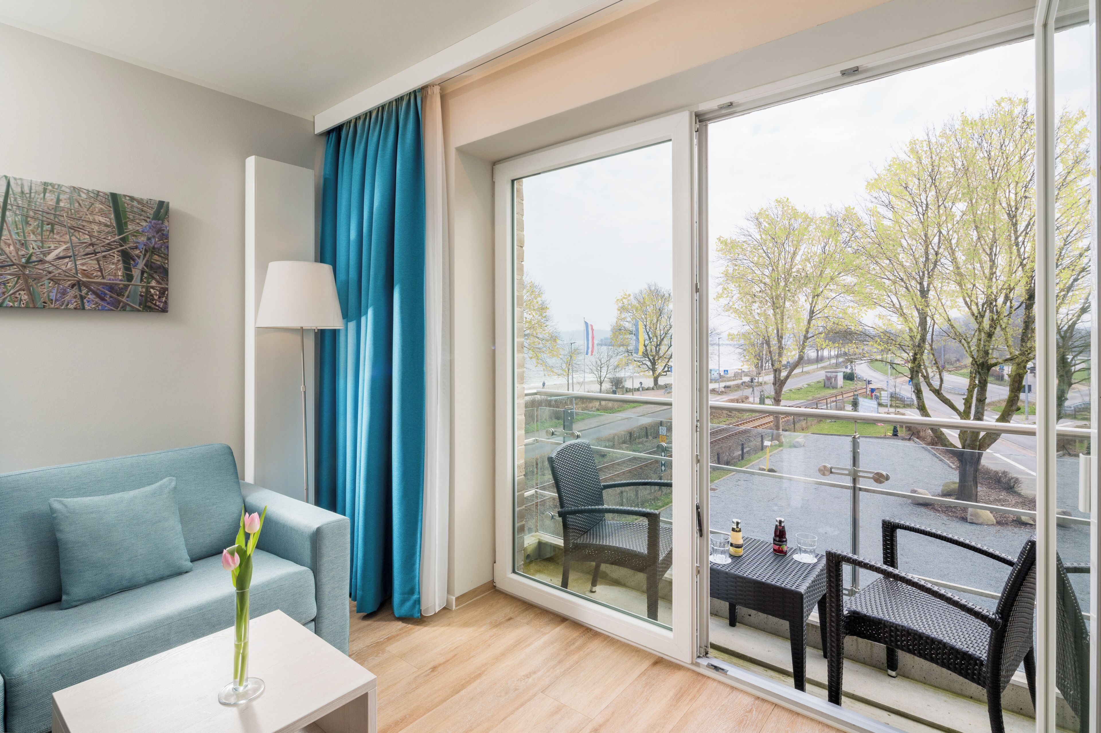 Una habitación moderna con sofá, una mesa con un jarrón de flores, un balcón con sillas y un ventanal con vistas a árboles y un paisaje urbano.