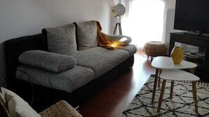 A modern living room with a grey sofa, a small white table, and a TV. There are wicker chairs and a patterned rug.